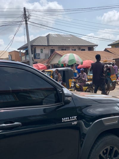 The image shows a black Toyota Hilux V6 parked in a busy street market in Ondo, Nigeria, located at latitude 7.251 and longitude 5.196. The street is lined with small shops and houses, some with solar panels on the roofs. Several people are going about their business, including a man in a blue shirt riding a motorcycle, a woman walking, and a man in a black shirt pushing a cart. The street is busy with traffic, including cars, motorcycles, and a yellow three-wheeled motorized vehicle called a "keke." The sky is blue with some white clouds. The image is taken from inside the parked Hilux, looking towards the front. The Hilux is positioned on the left side of the street, and the camera is aimed towards the center of the street, with the right side of the image partially obscured by the Hilux's body. The image is taken from the driver's side of the vehicle, looking towards the street. The car's door is open, and the front tire is visible. The street appears to be made of concrete, and the vehicles in the distance are all relatively small, suggesting a densely populated urban area.