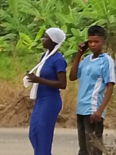 The image shows two young individuals standing outdoors during what appears to be daytime. The background is dominated by lush green foliage, possibly banana plants or similar large-leafed vegetation, suggesting a tropical or subtropical environment, or a rural/semi-urban setting.

On the left, a person, likely a young woman, is visible in profile facing left. She has dark skin and is wearing a white head covering or wrap, part of which hangs down over her shoulder and is held in her hands. She is dressed in a long-sleeved, royal blue dress that extends down her body. Her posture is relaxed, with her head slightly tilted up.

On the right, another person, likely a young man, is partially visible, facing slightly towards the left side of the frame but looking forward. He also has dark skin and is holding a dark-colored smartphone to his right ear, seemingly in a call. He has short, dark hair with a distinct haircut. He is wearing a light blue short-sleeved t-shirt with white stripes or panels on the sides and sleeves. The bottom part of his attire consists of dark pants.

The ground beneath them appears to be an unpaved road or dirt path, with some brown earthy tones visible. The overall image quality is somewhat soft or blurry, but the key details of the individuals and their actions are discernible. There is no visible text in the image.