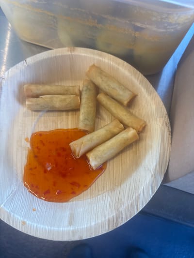 A close-up, top-down shot shows a light brown, round plate made of pressed palm leaves. On the plate are seven spring rolls, golden brown and fried. Some of the spring rolls are clustered together on the left side of the plate, while others are spread out towards the right. A pool of bright orange, viscous sweet chili sauce covers a portion of the right side of the plate, with a few stray droplets on the palm leaf. The background of the image is blurred, showing what appears to be a clear plastic container on the left and a dark blue surface on the right. The lighting suggests an indoor setting, possibly a food stall or restaurant. There is no visible text in the image. The scene depicts food ready to be served or consumed.