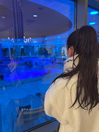 The image displays an interior perspective of an indoor aquatic center in Marcq-en-Barœul, France, viewed through a large glass partition. In the foreground, positioned with their back to the viewer, is an individual wearing a white, textured bathrobe. This person has long, dark hair tied into a high ponytail and is looking out through the window.
Beyond the glass, the midground is dominated by a spacious, irregularly shaped swimming pool filled with water intensely illuminated by an electric blue light. Approximately a dozen individuals are partially submerged in the pool, engaged in various leisure activities. On the poolside deck adjacent to the window, a light-colored slatted lounge chair is visible. The floor surrounding the pool consists of light-toned tiles.
In the background, the facility's modern architecture is evident, featuring a high, curved ceiling equipped with integrated blue lighting fixtures. A distant doorway reveals a solitary, indistinct human figure. The overall environment is characterized by cool blue illumination reflecting off surfaces, particularly the water.