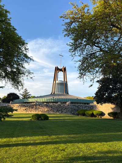 The image shows a low-angle view of a church, framed by trees, in Niles, United States of America. The church features a tall tower with a cross at its peak, set against a blue sky with wispy clouds. The building is constructed with a stone facade and a green roof. Manicured green lawns surround the building, with decorative bushes and trees nearby. Shadows cast by the trees streak across the grass.