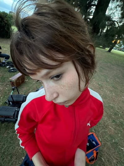 A person with brown hair and freckles poses close to the camera. They wear a red jacket with white stripes on the sleeves. The background consists of a grassy area with trees in the distance. Beside them is a blue and orange bag, as well as some camera equipment, including a box and two batteries. The shot is taken at an angle, making the background appear tilted.