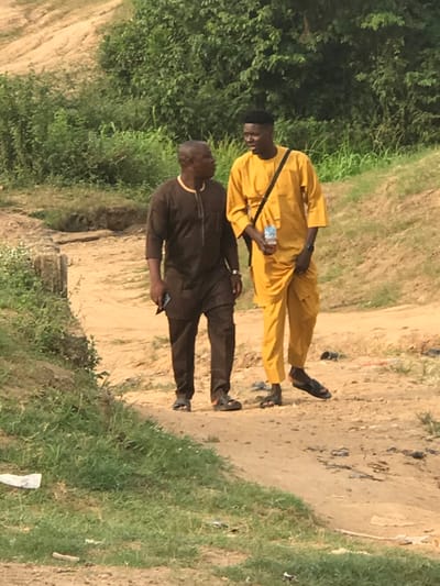 Two men in traditional attire walk along a sandy path. The man in front wears a bright yellow outfit with a black crossbody bag, and holds a water bottle. Behind him, the second man is dressed in a brown outfit and carries a phone. Both wear sandals. Lush green vegetation lines the path on the left and right. The background suggests a rural or semi-rural setting.