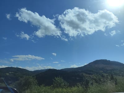 The image depicts a bright, sunny day with a clear blue sky. The upper portion of the image is dominated by a large, fluffy white cloud formation stretching horizontally across the frame. The sun is visible in the upper right corner, casting a bright glare and creating lens flare. Below the sky, a landscape of rolling, tree-covered hills is visible. The hills are a deep green, with some areas appearing darker due to shadows. The foreground consists of a grassy area with some trees and foliage, partially obscuring a car's side mirror and window on the lower left. The scene captures a natural, rural environment. Based on the provided context, this scene is located in Petrova, Romania.