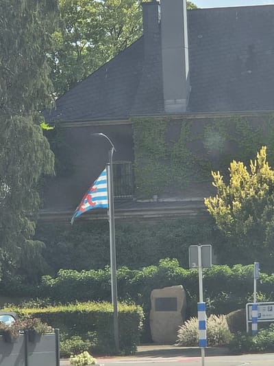 The image shows a flag pole with a flag that has blue and white horizontal stripes and a red lion with blue crowns and claws. This is the flag of Luxembourg. Behind the flag is a building covered in ivy, with a dark, sloped roof and a chimney. In the foreground, there is a stone monument with a dark plaque, a street sign, and a parking sign indicating "Op Acker". The scene is set outdoors, likely in a public area, with greenery and trees surrounding the monument and building. The lighting suggests it is daytime.