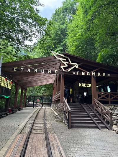 The image depicts the entrance to a train station in Guamka, Russia. The station building is constructed of dark wood, with a triangular roof and wooden support beams. Above the entrance, the words "ГУАМКА" are displayed in large, white, capital letters, framed by quotation marks. To the right, the word "КАССА" is also visible in yellow capital letters, indicating the ticket office. Below the main entrance, the word "СТАНЦИЯ" is written vertically in white capital letters on a wooden beam.

In the foreground, a railway track runs alongside a paved walkway. The track is made of gravel and wooden sleepers, with dark metal rails. The walkway is paved with light grey interlocking bricks. To the left of the building, lush green trees and foliage create a natural, forest-like setting. The sky is visible through the leaves, suggesting it is daytime.

Inside the station building, several people are visible. One person in a yellow shirt is standing near a counter, possibly interacting with a ticket seller. Another person in a dark top is also visible near the counter. A few individuals are seated on benches outside the building, waiting. In the distance, a black metal fence encloses part of the station area.

A digital display board is visible at the bottom of the image, showing rows of numbers and text, likely indicating train schedules or prices. The overall scene conveys a functional, yet rustic, train station environment nestled within a natural landscape. The presence of the railway, the station building, and people suggests that a train service is either operating or about to operate.