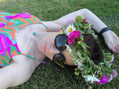 A person is lying on their back in a grassy field, outdoors. They are wearing a colorful swimsuit with a psychedelic pattern of oranges, pinks, purples, and greens. A delicate gold necklace with a turquoise clover pendant rests on their collarbone. Sunglasses with dark lenses are perched on their nose, and a flower crown made of daisies, pink carnations, clover, and various green foliage adorns their head. Their right arm is extended, and they are wearing a black smartwatch on their wrist. The sun appears to be shining, suggesting a daytime setting. The grass is green and well-maintained. The overall impression is one of relaxation and enjoyment of a summer day in a natural setting. The scene is likely peaceful and serene. No visible text is present in the image.