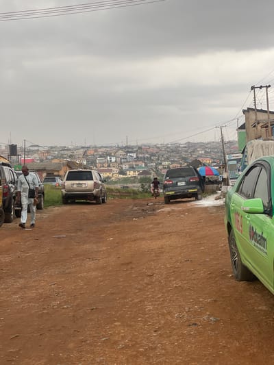 The image depicts a scene in Ojodu, Nigeria, under an overcast sky. A row of parked vehicles, including a green "ODUAM" branded car, a dark SUV (license plate JJJ207JN), and a white minivan, line a muddy, reddish-brown roadside. Two individuals, dressed in white traditional attire, stand near the front of the row. In the background, a densely populated urban landscape of primarily low-rise buildings stretches into the distance. Power lines crisscross overhead. A billboard with "Montepoin" is visible near the green car.