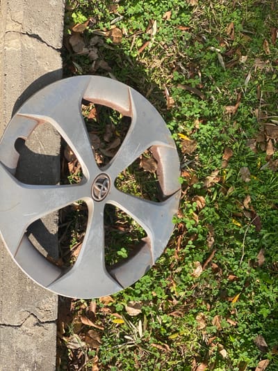 A silver-colored car hubcap with a five-spoke design lies on the ground in Jaljulia, Israel. The hubcap, showing signs of wear with scratches and reddish-brown discoloration along the inner edges of its spokes, prominently features the circular Toyota logo in its center. It rests partially on a rough, cracked concrete curb and partially on a patch of lush green grass mixed with clover-like plants. Numerous dried, brown leaves are scattered across the grassy area. The scene is brightly illuminated by direct sunlight, casting distinct shadows from the hubcap, indicating a clear and sunny day. No individuals or specific activities are depicted in the image.
