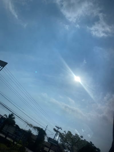 The image shows a bright, sunny sky with scattered clouds, possibly during the day in Sungai Lakum, Indonesia. The sun is visible, emitting strong rays that create a lens flare effect. Several power lines crisscross the frame, suggesting an urban or semi-urban environment. Below the sky, there are glimpses of trees, foliage, and what appears to be part of a building or structure, likely near a roadside given the presence of utility poles and wires. The perspective suggests the photo was taken from a moving vehicle or an elevated position.