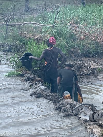 Two individuals stand by a muddy riverbank. One, wearing a maroon head covering, black shirt, and orange undershirt, bends while holding a black bucket. The other person stands with a tool and is clad in a black shirt with a yellow undershirt. Mud, grass, and various plants surround the riverbank. Water flows in the river.