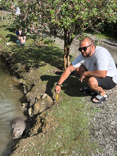 A man wearing sunglasses, a white t-shirt, and black shorts squats on the ground near a body of water, holding a yellow object in his hand. A nutria is partially submerged in the water near the bank, facing the man. In the background, several people are visible standing under a tree with red and white flowers. The environment appears to be a park or natural area with grass, trees, and a gravel path. The image does not provide any specific information about the location being in Semenovka, Russia.