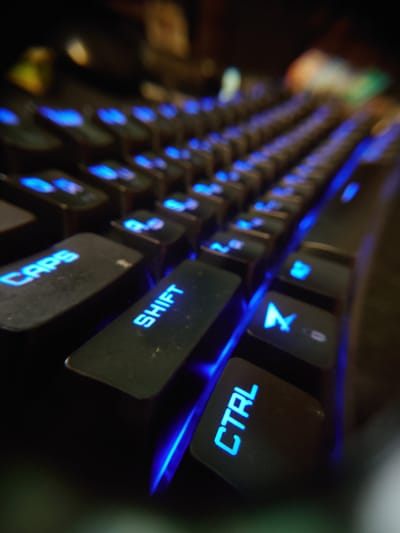This is a close-up, low-angle shot of a black mechanical keyboard with blue backlighting. The focus is on the keys, with some appearing in sharp focus while others are blurred due to the shallow depth of field. Visible keys include "CAPS", "SHIFT", and "CTRL", all illuminated by the blue LEDs. The overall scene is dark, with the blue light from the keyboard being the primary light source. The background is out of focus, showing blurred shapes and colors that suggest a dimly lit room. There is no indication of the city of Sokolohirsk, Ukraine, in the image.