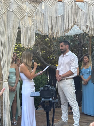 An outdoor ceremony is depicted on a wooden platform beneath a large macrame-decorated canopy in Beit Yehoshua, Israel. A woman with long blonde hair, wearing a white sleeveless floor-length dress, stands centrally holding a black microphone in her right hand and a white sheet of paper in her left. She is positioned to the left of a black professional camera mounted on a black tripod, which faces a man with dark hair and a beard. This man, dressed in a white long-sleeved shirt, light beige trousers, and a brown belt, stands with his hands clasped in front of him, looking towards the speaking woman. To the left of the speaking woman, a second blonde woman in a light green strapless dress stands with her hands clasped. To the right and slightly behind the man, a third blonde woman in a light blue ruffled-sleeve dress also stands with her hands clasped. Dense green foliage from trees and bushes forms the background.