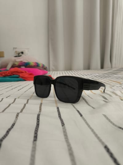 A pair of black sunglasses with dark lenses rests on a white bedsheet with thin, vertical gray and brown stripes. The sunglasses are positioned in the foreground, slightly off-center to the right. In the blurred background, a pile of colorful clothing, including pink, orange, and teal fabrics, is visible on the bed, along with a fluffy white pillow. A wall with a white outlet and a light switch is also discernible behind the clothing. The scene appears to be indoors, likely in a bedroom, possibly in General Trias, Philippines.