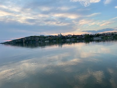 The image shows a body of water with reflections of clouds in the sky. On the horizon is the city of Harsova, Romania. The city is situated on a hill with several buildings and structures visible. A large church with golden domes is prominent in the skyline. There are trees and vegetation along the shoreline, with several boats docked along the riverbank. There appears to be a monument or statue on the hill to the left of the skyline.