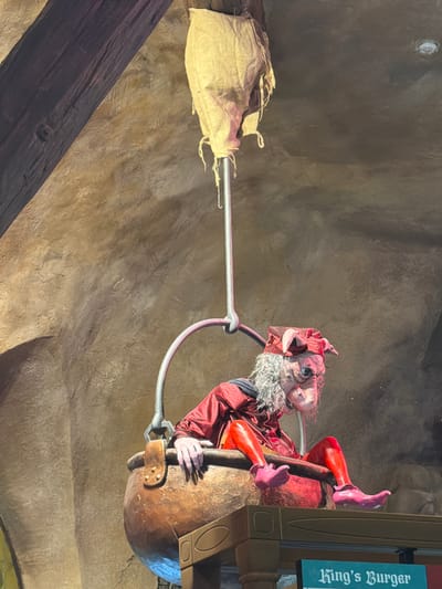 The image shows a puppet resembling a goblin with grey hair, a red tunic, and pink shoes, sitting in a cauldron that is suspended by a metal rod from a burlap sack. The puppet is positioned in front of a textured, cave-like wall. Below the cauldron, a sign reads "King's Burger". This scene appears to be part of an attraction or themed restaurant, likely in the city of Castelnuovo del Garda, Italy, which is known for its theme parks.