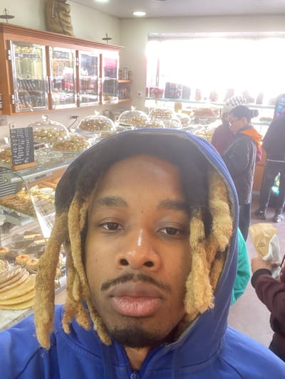 This image depicts the interior of a bakery or pastry shop located in Le Pré-Saint-Gervais, France.

In the immediate foreground, a Black male individual is captured in a close-up, selfie-style photograph. He has a close-cropped beard and mustache, and shoulder-length dreadlocks with prominent blonde tips. He is wearing a blue hooded sweatshirt, with the hood pulled over his head, and gazes directly at the camera with a neutral expression.

The midground and background reveal the shop's interior. Numerous baked goods, including various pastries, cookies, and layered sweets, are displayed in glass cases and under clear glass cloches. A small blackboard sign with white handwritten text, presumably a menu or price list, is visible on the left side, resting near a display case. Above the display cases, a wooden cabinet with multiple glass-paneled doors is mounted on the wall, holding various packaged items. A large, light-colored burlap sack rests on top of this cabinet. Overhead lighting fixtures are present.

Several other individuals are visible in the background. On the right, a person wearing a dark jacket with an orange lining and eyeglasses is observed looking downwards. Another person, partially obscured, wears a white beanie and a maroon top. Further back, a third individual holds a light-colored paper bag. The far background features a large window or glass storefront, through which bright, possibly daylight, is intensely visible, obscuring exterior details.

The scene suggests a bustling commercial environment, with the foreground subject actively taking a photograph and background individuals appearing to be customers browsing or engaged in transactions.