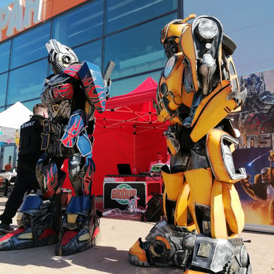 Two individuals in elaborate Transformers costumes, one resembling Optimus Prime (blue, red, and grey with flame decals) and the other Bumblebee (yellow and dark grey), stand on light-colored pavement in front of the Globus Hypermarket in Трубачеевка, Russia. The building features a large facade of blue-tinted glass windows reflecting the clear blue sky, with white block letters spelling "PAH" or "БАН" visible on an orange upper section.
Behind the Optimus Prime costume, a man wearing dark pants and a black vest with "SWAT" in white letters on the back stands facing away from the camera, near a white pop-up tent adorned with colorful bunting. Between the two Transformer costumes, under a red pop-up tent, a man with a light beard is seated at a table with a laptop and a water bottle, operating what appears to be sound or event equipment. A black case bearing the "Globus" logo and various cables are visible on the ground near his setup. To the far right, behind the Bumblebee costume, a large banner or poster featuring other Transformers characters and the "TRANSFORMERS" logo is displayed. The scene is brightly lit by direct sunlight, casting distinct shadows on the pavement, indicating a daytime outdoor event.