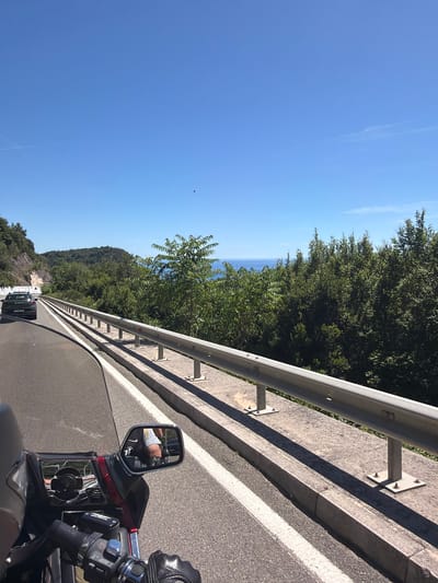 The image depicts a view from a motorcycle traveling on a coastal road in Brežine, Montenegro. The road curves to the left, with a metal guardrail separating the traffic lane from a steep drop-off. Lush green trees and vegetation line the right side of the road, leading to a glimpse of the blue Adriatic Sea in the distance. A dark-colored car is visible ahead on the left lane. The sky is a clear, bright blue, with a few faint wisps of clouds. The motorcycle's windshield and handlebars are visible in the foreground, with a side mirror reflecting the rider's legs.