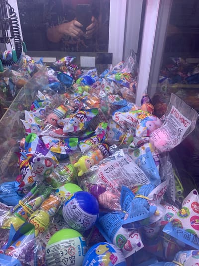 The image displays a close-up view of the interior of a claw machine filled with various small, individually packaged toys. The toys are predominantly colorful and appear to be made of soft materials. Many of them are round or animal-shaped. The packaging is mostly transparent plastic, with colorful printed designs. Some packages have text and symbols, including "COOL THINGS" and "CE" markings. The lighting inside the machine appears to be bright and artificial, casting some shadows. The reflection on the glass in the upper part of the image shows a person holding a smartphone, possibly taking the photograph, and a dark, patterned garment. The scene is consistent with a common arcade or entertainment center setting. The location is identified as Sinalunga, Italy.