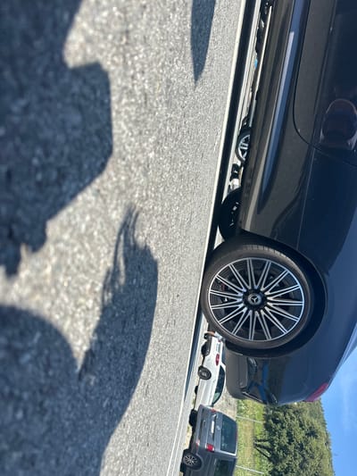 The image shows a parking lot in Varazze, Italy. The angle is low, focusing on the asphalt and the shadows cast by a person taking the picture.
In the background, there are three vehicles:
1. A dark-colored coupe or sedan, with a distinctive multi-spoke wheel design, that seems to be a Mercedes-Benz.
2. A white station wagon of the same brand.
3. A dark gray Volkswagen Transporter van.
The location appears to be a sunny, outdoor parking area. Green vegetation is visible in the distance.