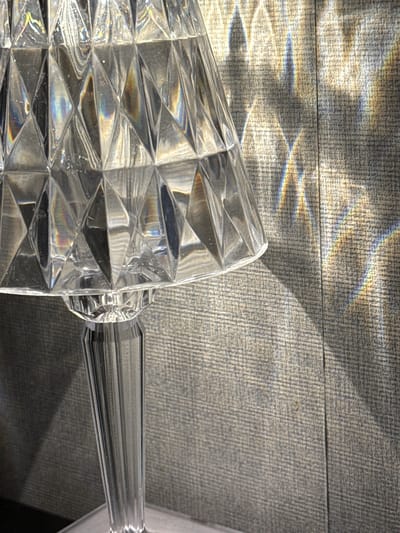 A crystal lamp with a faceted shade and a fluted stem stands on a surface, casting prismatic light patterns onto a textured, grey wall. The lamp appears to be made of clear glass or acrylic, with a diamond-cut pattern on the shade that refracts light into rainbow hues. The wall behind the lamp has a subtle, woven texture and a vertical seam running through it. The lighting suggests it is daytime, with light coming from an unseen source, possibly a window, that is illuminating the lamp and creating the light projections on the wall. The scene is static, with no people or actions depicted. The overall impression is one of elegant, decorative lighting. The location in Gwangju, South Korea, is not discernible from the image itself.