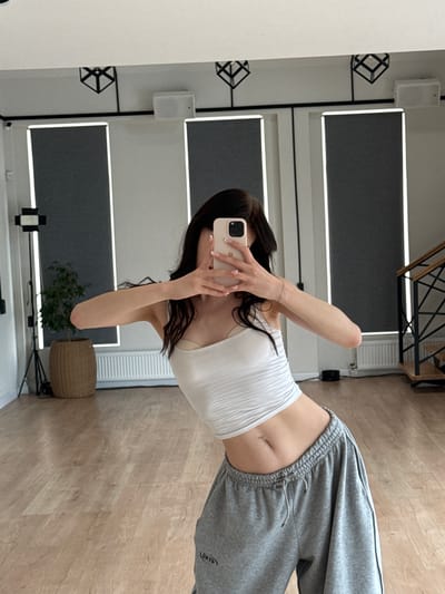 The image shows a young woman taking a selfie in a well-lit room with a light wood floor. She is wearing a white strapless top and grey sweatpants. She has long dark hair and a belly button piercing. The room appears to be a modern space, possibly a home or studio, with sleek minimalist decor. In the background, there are large rectangular panels with thin white light strips, and on the left wall, there are cubic decorative shelves and a white speaker. A woven basket and a plant are visible in the corner of the room. The overall impression is one of casual relaxation and personal style. The lighting suggests it is daytime. There is visible text on the sweatpants which appears to read "CYBER".