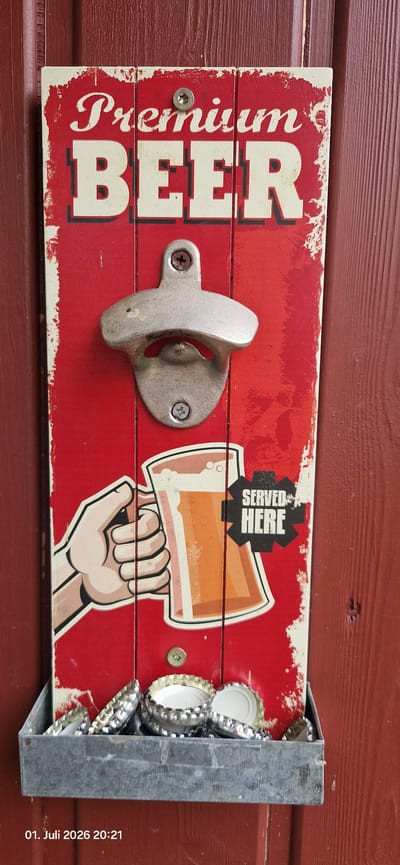 This media file shows a decorative wooden beer bottle opener with a built-in bottle cap catcher. The opener is mounted on a reddish-brown wooden surface, likely an exterior wall or fence, common in a rural setting. The wooden board has a distressed red finish with white accents, featuring large white lettering that reads "Premium BEER". To the right of the bottle opener, there's a graphic illustration of a hand holding a glass of beer, with the text "SERVED HERE" superimposed on the glass. The bottle cap catcher is made of galvanized metal and is filled with numerous silver and white bottle caps. The scene appears to be outdoors, given the lighting and the wooden background. The timestamp "01. Juli 2026 20:21" is visible in the bottom right corner, indicating it was captured on July 1, 2026, at 8:21 PM. The setting is identified as Hummeltal, Germany. There are no people visible in the image. The overall mood is casual and evokes a sense of a beer-drinking establishment or a personal bar area.