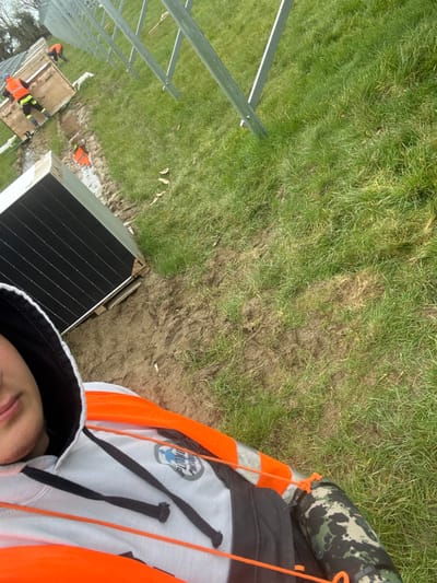 The scene depicts an outdoor construction site at Baldurgan Hill, Ireland, involved in the installation of a solar energy array. In the foreground, a person is partially visible, wearing a grey hooded sweatshirt with a logo and an orange high-visibility vest. Orange lanyards are visible across their chest, and a camouflage-patterned cylindrical object is attached to their right side. To the left, a stack of black solar panels is resting on a wooden pallet. A muddy, disturbed strip of ground traverses the midground, separating the foreground from the main work area. Two additional workers are present in the midground; one, wearing an orange high-visibility top, is positioned near several large wooden crates, some labeled "SOAR 2.0," while another worker, also in an orange high-visibility top, is bending over further back. A line of partially installed metallic (likely galvanized steel) solar panel mounting frames extends diagonally from the mid-left towards the far right. The environment consists of a grassy field with significant patches of disturbed earth and mud. Overcast conditions are indicated by the diffused lighting.