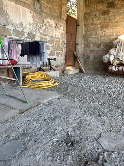 In Jrarbi, Armenia, this image captures a rustic outdoor scene. A clothesline hangs with several items of clothing, including a pink shirt and dark t-shirts. A coiled yellow garden hose lies on the ground next to a grey concrete surface. The background features a brick wall and a weathered brown door. The ground is a mix of concrete and gravel.