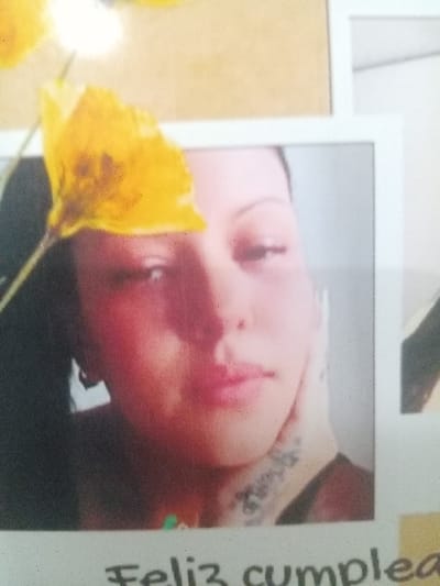A close-up image captures a person's face in a framed photograph. A yellow flower is positioned in the lower left corner, partially obscuring the person's face. The person has fair skin, pink lips, and appears to have a tattoo on their upper shoulder/neck area. Text is visible on the right edge of the frame that reads "Feliz cumple". The image is slightly blurred.