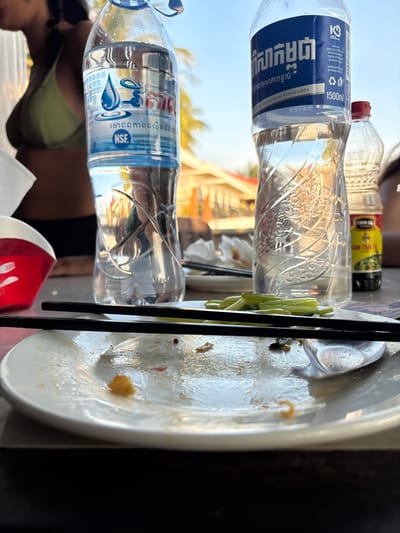 The image depicts a post-meal scene at an outdoor dining establishment in Sok San Village, Cambodia, under bright daylight. In the foreground, a white, round plate rests on a dark surface, displaying sparse food residue including orange crumbs, red specks, and dark smears. Two black chopsticks are positioned horizontally across the plate, alongside a partially visible silver spoon on the right. Directly behind the plate, a small white dish contains a pile of green, elongated vegetables. To the left, a clear plastic bottle of "Phnom Srok" brand bottled water, featuring a blue label with Khmer script and an NSF certification mark, is visible. To the right, a larger 1500ml clear plastic bottle of "Sok San" brand bottled water, also with a blue label in Khmer script indicating adherence to European standards, stands prominently. Adjacent to it is a small glass bottle with a red cap and a yellow "CHIN-SU Tam Thai Tu" label, containing a dark liquid, likely chili sauce or soy sauce. In the blurred mid-background to the left, the upper torso of an individual, partially visible, is wearing a green ribbed bikini top and dark bottoms. The distant background shows palm trees and light-colored building structures under a clear blue sky, indicating an outdoor or semi-outdoor dining setting.