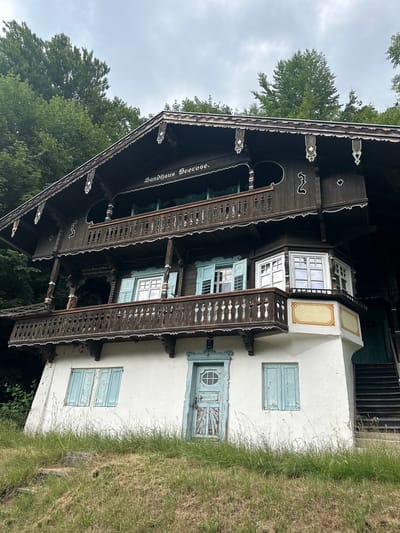 The image shows a view of the "Seerose" guesthouse in Kufstein, Austria, a picturesque town nestled in the Tyrolean Alps. The guesthouse is a traditional Austrian wooden house, characterized by its dark wood facade adorned with intricate carvings and white decorative trim. It features multiple balconies with wooden railings, and light blue shutters on some of the windows. The lower portion of the house is plastered in white, with a light blue door and a window with similar blue shutters. The guesthouse is surrounded by lush green trees, suggesting a natural, possibly mountainous, setting. The sky is overcast, contributing to a somewhat subdued lighting. The foreground is covered in green grass, with a few stone steps leading up to the house, indicating a slightly inclined terrain. There are no people visible in the image.