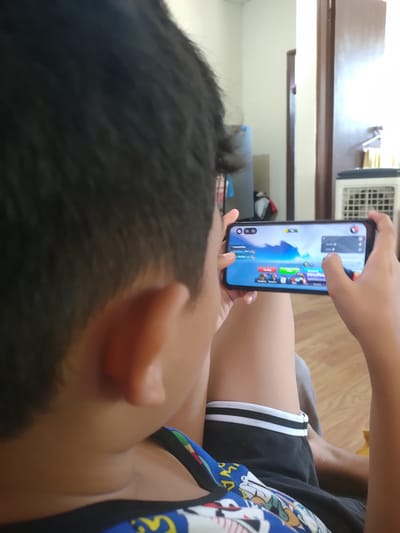An over-the-shoulder view shows a young person playing a mobile game. The game displayed on the phone screen appears to be a battle royale or similar action game. The individual is holding the phone horizontally with both hands. The background suggests an indoor setting, possibly a residential room. Elements visible include wooden flooring, a doorway, and a white air conditioning unit. The overall scene is casually lit, indicating it might be daytime. The location is identified as San Pascual, Philippines.