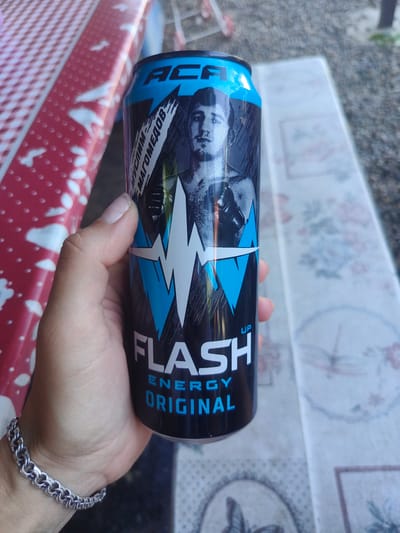 A hand holds a can of "FLASH ENERGY ORIGINAL" drink. The can has a black background with blue and white graphics, including a lightning bolt design and a portrait of a man. The can is positioned in front of a red and white checkered surface and a blurred background. The scene appears to be outdoors, possibly on a table. There is no discernible information within the image to link it to the city of Staronizhestebliyevskaya, Russia.