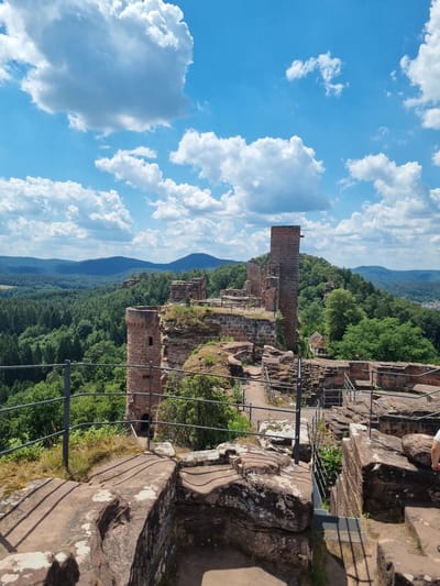 This is a landscape photograph taken from a high vantage point, possibly a castle ruin, overlooking a vast, tree-covered valley. The sky is a vibrant blue with scattered white clouds. The foreground features weathered stone structures, remnants of a historical building, with metal railings and staircases suggesting a tourist path. The ruins are made of rough-hewn stone blocks.
In the mid-ground, the scene transitions to a dense, green forest that carpets the rolling hills and mountains stretching into the distance. The landscape evokes a sense of history and natural beauty. The lighting suggests it is daytime, likely a sunny day given the bright blue sky and clear visibility. The overall mood is serene and picturesque, highlighting the juxtaposition of ancient architecture and the enduring power of nature. There is a hint of a person's leg and foot at the top edge of the frame, wearing a red shoe, indicating human presence and exploration of the site. The location is identified as Dahn, Germany, known for its unique rock formations and castle ruins. There is no visible text in the image.