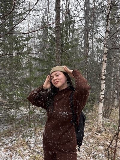 A young woman stands in a snow-covered forest in Kachkanar, Russia, smiling and posing for a photo. She wears a brown sweater dress and a light-colored knit hat, shielding her eyes with her hand. Bare trees and evergreens surround her, with a birch tree visible in the background. Light snow falls. A black bag is slung over her shoulder.