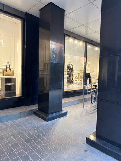 The image shows the exterior of a boutique called "belle lingerie" in Novaya Usman, Russia. The store features a large display window with mannequins dressed in clothing, and a beige handbag displayed on a black tiered stand. The shop's logo, "belle" and "boutique lingerie," is visible on the window. Outside the store, there is a bicycle parked near a metal railing, and the entrance area is paved with stone tiles. The building has a modern architectural style with dark stone pillars and a tiled ceiling.