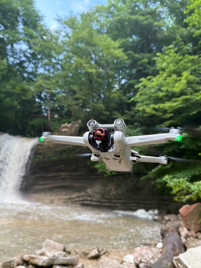 An aerial drone, specifically a DJI Mini 3 Pro, hovers in mid-air in front of a waterfall. The drone is a light gray color and is positioned in the foreground, occupying a significant portion of the frame. Its propellers are visible, with the front right propeller illuminated by a green light. The drone's camera gimbal is angled downwards, and a small, reddish object is visible within the gimbal.

In the background, a waterfall cascades down a rocky cliff face. The water is white and foamy, contrasting with the darker, layered rock. A river flows at the base of the waterfall, appearing murky and shallow. The riverbanks are lined with rocks and pebbles.

The scene is set within a lush, green environment. Tall trees with dense foliage form a backdrop behind the waterfall and drone. Sunlight filters through the leaves, creating dappled light and bokeh effects, especially in the upper portions of the image. The sky is visible through the trees, appearing light blue with some white clouds. The overall impression is of a natural, remote location. The image does not contain any people.