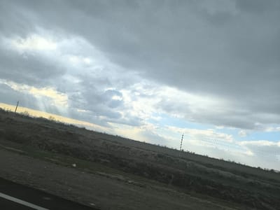 The image displays a tilted landscape captured in Pakhtakor, Uzbekistan, under a cloudy sky. The upper frame is dominated by heavy grey clouds, through which crepuscular rays of sunlight penetrate in the upper left, illuminating the distant horizon with a yellow hue. Sections of blue sky are visible in the center-right and lower right, with a distinct circular void or formation present within the clouds near the image center. The foreground and mid-ground consist of dark brown earth, indicative of tilled fields or barren land, interspersed with patches of green vegetation. The terrain shows a gentle slope from the foreground towards the horizon. A dark utility pole with cross-arms is positioned on the left side of the image, while a lighter-colored, segmented pole with horizontal utility wires extends across the right mid-ground. A sparse line of trees or bushes marks the distant horizon. The immediate foreground features a dark, linear band, likely the interior of a vehicle or a road surface, with a diagonal white line. No individuals or specific activities are visible within the scene.
