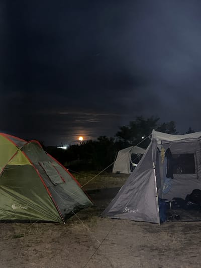 The image depicts a campsite at night, illuminated by a bright full moon visible in the dark sky. Two tents are prominently featured: a green and gray tent on the left, and a larger gray tent on the right, which appears to be open, revealing some items inside. The surrounding environment consists of dry ground with scattered vegetation. In the background, trees and distant lights are visible, suggesting a possible proximity to an urban or semi-urban area. The sky is partly cloudy, with the moon shining through. The scene is consistent with a campsite in the vicinity of the village of Kolodezny, Russia, given the natural and lightly developed landscape.