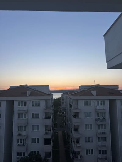 The image depicts a view of apartment buildings in Çanakkale Merkez, Turkey, during either sunrise or sunset. The sky is a gradient of light blue, transitioning to warmer hues of orange and pink near the horizon. In the foreground, two parallel apartment buildings are visible, characterized by their white facades, multiple floors with numerous windows, and balconies. The buildings have red-tiled roofs. A body of water, likely the Dardanelles strait, is visible in the distance, behind a line of trees. The street level between the apartment buildings is visible and appears to be a road with some parked motorcycles. The overall scene is quiet and residential, capturing a moment of transition in the day.