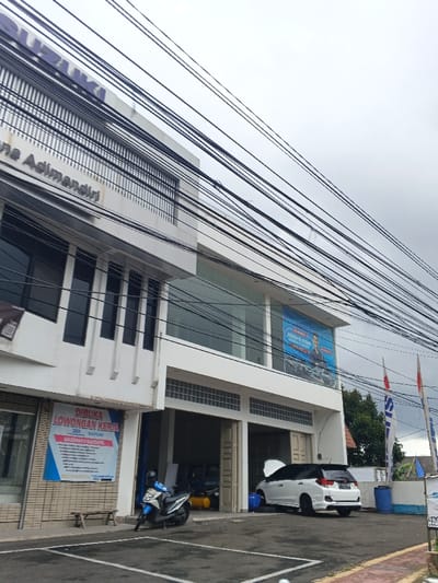 The image displays the exterior of a two-story white commercial building, identified by "SUZUKI Adimandiri" signage near the top left. Numerous black utility lines traverse the upper portion of the frame against an overcast sky.
On the ground floor, multiple open bays are visible, suggesting a service center or showroom. A white car with a black roof spoiler is parked partially inside the rightmost bay, its hood open, indicating maintenance or inspection. A blue plastic drum is positioned next to this vehicle. Inside the same bay, a yellow air compressor is visible. To the left, a light blue and white scooter is parked in front of another bay.
A vertical banner on the building's left side reads "DIBUKA LOWONGAN KERJA" (Job Vacancy), listing positions for "SALESMAN & SALESFORCE" for "DEA SUZUKI". Below this banner, a simple wooden bench rests on the pavement. On the upper floor, a large blue promotional banner or sign featuring two individuals and vehicles is displayed behind a window. Two flagpoles with flags, one red and white, and another blue with a Suzuki logo, are situated to the right of the building. The ground area is paved with parking lines.
Regarding the specified location, the visible branding "Suzuki Adimandiri" is typically associated with Suzuki dealerships operating in Indonesia. There are no visual cues within the image that definitively confirm its location in Palayan, Philippines; the branding instead suggests an Indonesian context.
