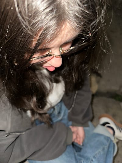 A low-angle, slightly out-of-focus image showing a person with dark hair and glasses lying down next to a black and white dog. The person is wearing a gray hoodie and blue jeans, and has on white sneakers with tan soles. The background appears to be a dimly lit outdoor area, possibly in a city, but not enough detail is visible to confirm the location or specifics of Agarak, Armenia. The person's face is partially visible, showing a light skin tone. The dog appears to be resting or sleeping.