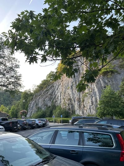 A parking lot is situated at the base of a large, gray, rocky cliff face in Durbuy, Belgium. The cliff is covered in some vegetation, with trees growing at its base and on ledges. The sky is bright and slightly overcast, with a faint contrail visible. In the foreground, the tops and hoods of several parked cars are visible, including a dark-colored SUV and a dark pickup truck. The trees in the foreground, with their green leaves, partially obscure the view of the cliff. In the background, more trees line the area behind the parking lot. A lamppost with a flower basket hangs from it is visible in the mid-ground. The scene appears to be a scenic spot frequented by visitors.