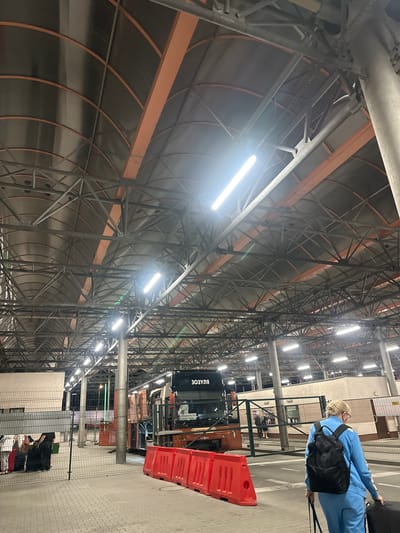 The image shows a bus station in Hrebenne, Poland. The station has a large, arched, metal roof supported by orange beams and grey pillars. Fluorescent lights are visible on the ceiling, illuminating the space. In the foreground, a woman in a blue tracksuit with a black backpack is walking away from the camera, pulling a black suitcase. To the left, a metal fence encloses an area where several pieces of luggage are gathered. Behind the fence, a bus with the word "ЗОЗУЛЯ" in Cyrillic on its side is parked. Red plastic barriers are placed on the ground in front of the bus. In the background, there are buildings and more fencing, suggesting a border crossing. The ground is paved with grey interlocking bricks.