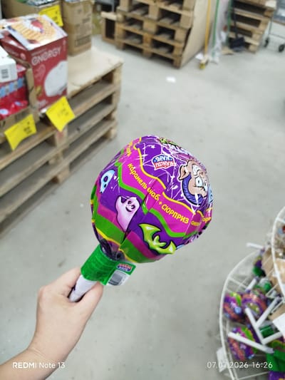 A hand holds a large, round lollipop with purple, green, and pink wrappers depicting cartoon characters, including a ghost and a boy with bug eyes. The lollipop wrapper has Russian text. In the background, shelves stacked with boxes and palettes are visible, suggesting a retail or warehouse environment. The floor is concrete. A basket of smaller lollipops is also in the background. The timestamp "07.07.2026 16:26" is present in the lower right corner. The location is identified as Nizhny Bestyakh, Russia.
