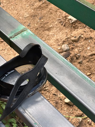 A pair of black swimming goggles rests on a weathered metal gate with green paint. The ground below is sandy with small rocks and debris. The goggles have water droplets on their straps and lenses, suggesting recent use. The gate's metal beams are worn and show signs of welding. Some green vegetation is visible at the bottom right corner.