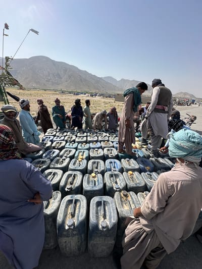 The image shows a large group of people gathered in what appears to be a rural or mountainous area in Unknown, Pakistan, suggested by the landscape and traditional clothing worn by the individuals. The focal point is a substantial pile of stacked blue plastic containers, likely jerrycans, with yellow caps. The people are standing around and on top of the containers. Many are dressed in traditional shalwar kameez and head coverings typical of the region. The background features a vast open space with mountains. The sky is clear, suggesting a sunny day. The image captures a scene that could represent a distribution point, possibly for water or fuel, or could simply be a storage location.